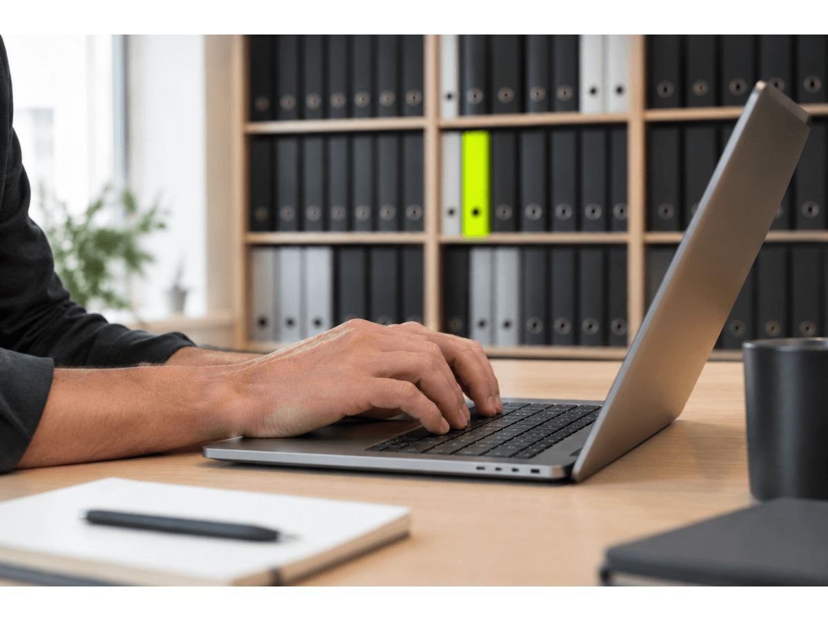 Hände tippen an einem Laptop im Büro, im Hintergrund ein Regal mit Ordnern